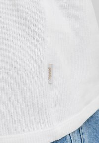 Gros plan sur un tissu blanc texturé avec une petite étiquette beige indiquant "Originals", porté avec un jean en denim bleu.