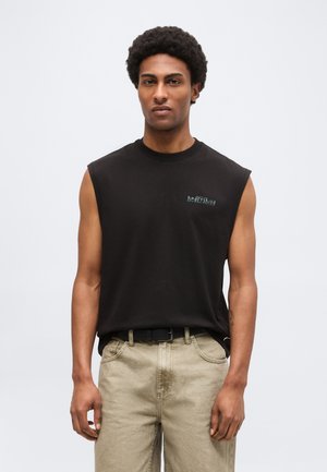 Homme aux cheveux bouclés portant un débardeur noir avec un texte sur la poitrine et un pantalon beige sur un fond gris uni.