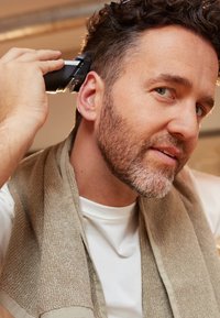 Zwarte haartrimmer gehouden nabij de zijkant van een man zijn hoofd. Hij draagt een wit T-shirt, met een lichtbeige handdoek over zijn schouders gedrapeerd.