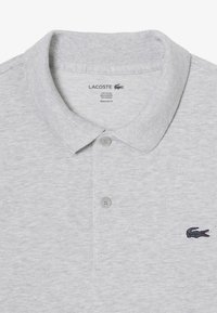 Ljusgrå polo skjorta i bomullsblandning, med klassisk krage, två knappar och en mörk broderad krokodil-logotyp på vänster sida.