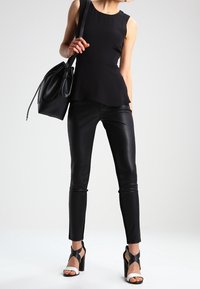 Haut péplum noir associé à un pantalon en cuir noir et des sandales à talons noires avec des brides blanches. Modèle tenant un sac en cuir noir.