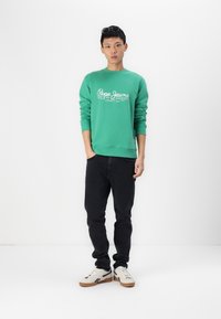 Sudadera verde con el logo blanco de "Pepe Jeans", combinada con unos vaqueros negros y zapatillas beige. El modelo está de pie con las manos en los bolsillos contra un fondo blanco.
