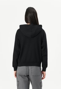 Woman with long dark hair wearing a black hooded sweatshirt and gray cargo pants, facing away against a plain white background.