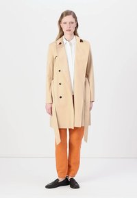 Manteau beige avec boutons noirs, taille ceinturée et col cranté. Le modèle porte un pantalon orange et des chaussures noires, avec une chemise blanche en dessous.