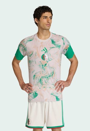 Mand står med hænderne i siden iført en pink og grøn Algeriet fodboldtrøje og hvide shorts med Algeriets emblem.