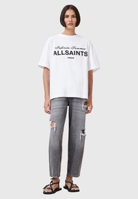 Witte oversized t-shirt met zwarte tekst "Infinite Summer ALLSAINTS LONDON," gecombineerd met versleten grijze jeans en zwarte sandalen.