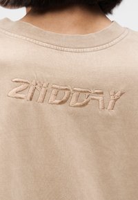 T-shirt en coton beige avec un col rond, présentant une broderie en relief du texte "2ND DAY" au dos. Texture lisse, design simple.