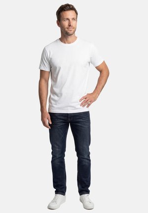 Mann trägt ein einfaches weißes T-Shirt, dunkelblaue Jeans und weiße Turnschuhe, steht mit einer Hand in der Hüfte vor weißem Hintergrund.