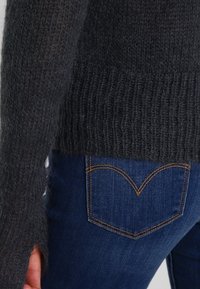 Pull en maille gris foncé avec une texture lâche et un ourlet côtelé, associé à un jean en denim bleu présentant des coutures distinctives sur les poches arrière.