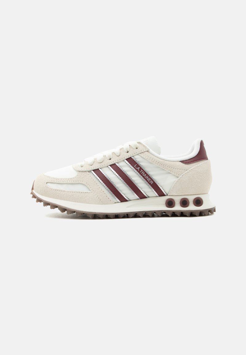 Baskets Adidas LA Trainer blanches et beiges avec des bandes bordeaux, un détail au talon et une semelle marron texturée, orientées vers la droite sur un fond gris clair.