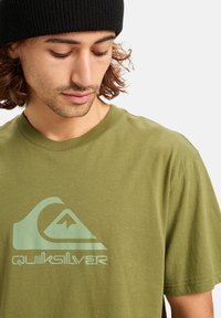 Olivově zelené bavlněné tričko s velkým světle zeleným logem Quiksilver. Design zahrnuje grafiku vlny a hory. Styl s kulatým výstřihem.