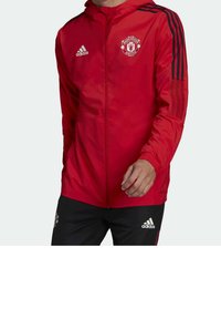 Chaqueta roja con cremallera y logo del Manchester United, con acentos de franjas negras en las mangas y puños elásticos. El material es ligero y brillante.