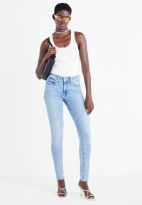 Jeans ajustados azul claro de cintura alta, camiseta blanca de tirantes con tejido acanalado y bolso negro cruzado. Gafas de sol amarillas y zapatos de tacón con correas completan el conjunto.