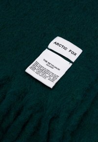 Türkisfarbener Wollschal mit fransigen Rändern, versehen mit zwei weißen Stoffetiketten, die den Markennamen "ARCTIC FOX" und Pflegehinweise anzeigen.