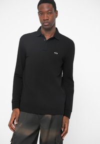 Camisa de polo preta de manga longa com colarinho, apresentando um pequeno logótipo branco no lado esquerdo do peito. Feita de um tecido liso, com um design minimalista.