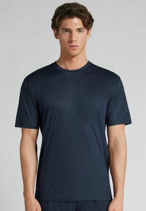 Joven con cabello castaño que lleva una camisa azul marino de manga corta con un bolsillo en el pecho, de pie frente a un fondo liso de color gris claro.