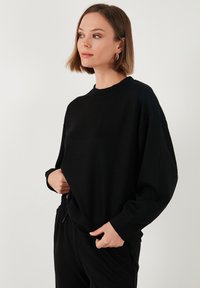 Felpa nera con scollo a costine, vestibilità ampia e maniche lunghe. Il tessuto sembra morbido con una texture liscia.