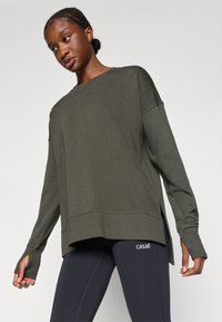 Sudadera verde oscuro con un corte holgado, mangas largas, aberturas laterales y agujeros para los pulgares. Presenta detalles de costura sutiles y un logotipo en las mallas.