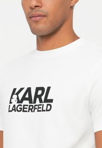 Valge puuvillane T-särk lühikeste varrukatega, mille esiküljel on julged mustad "KARL LAGERFELD" tekst ja siluett graafika.
