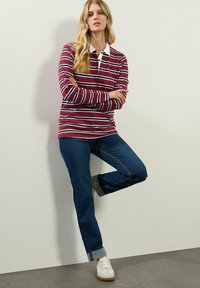Czerwono-biały w paski, długi rękaw polo z białym kołnierzykiem, w zestawie z niebieskimi dżinsami i białymi sneakersami. Model stoi pod ścianą.