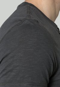 Gros plan sur l'épaule d'une personne portant une chemise en tissu texturé gris foncé avec des coutures visibles.