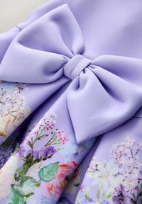 Levandų spalvos audinio kaspinėlis ant gėlių raštuoto audinio, kuriame yra rožinių, baltų, violetinių gėlių ir žalių lapų švelnių pastelinių spalvų.