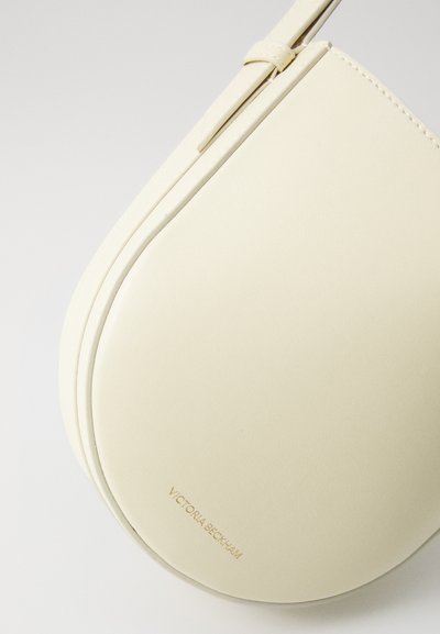 Krēmkrāsas ādas rokassoma ar minimālistisku ovālu formu, ar smalku šuvju un debosētu zīmola nosaukumu apakšā.