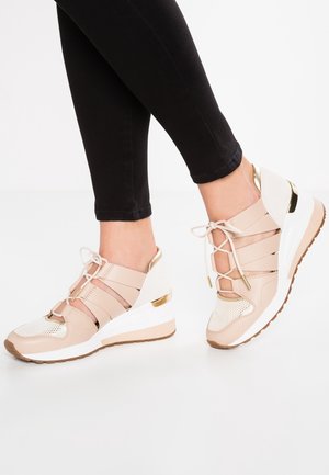 Nohy obuté v béžovo-bílých a zlatých wedge teniskách se šňůrkami a průstřihy po stranách, v kombinaci s černými přiléhavými kalhotami.