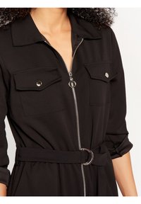 Black belted jumpsuit featuring a zip front, two chest pockets, and rolled-up sleeves. Made from smooth fabric with a structured design.