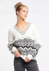 Fehér kötött pulóver fekete geometrikus mintával, V nyakú, rojtos részletekkel az alján és az ujjak végén, texturált keveréke a zsinóros és a sima kötésnek.
