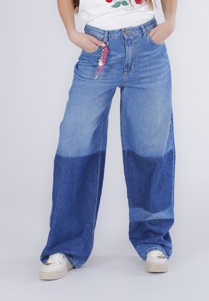 Mujer con vaqueros azules de dos tonos y pierna ancha con bolsillos, zapatillas blancas y una camisa blanca con un diseño floral, manos en los bolsillos.