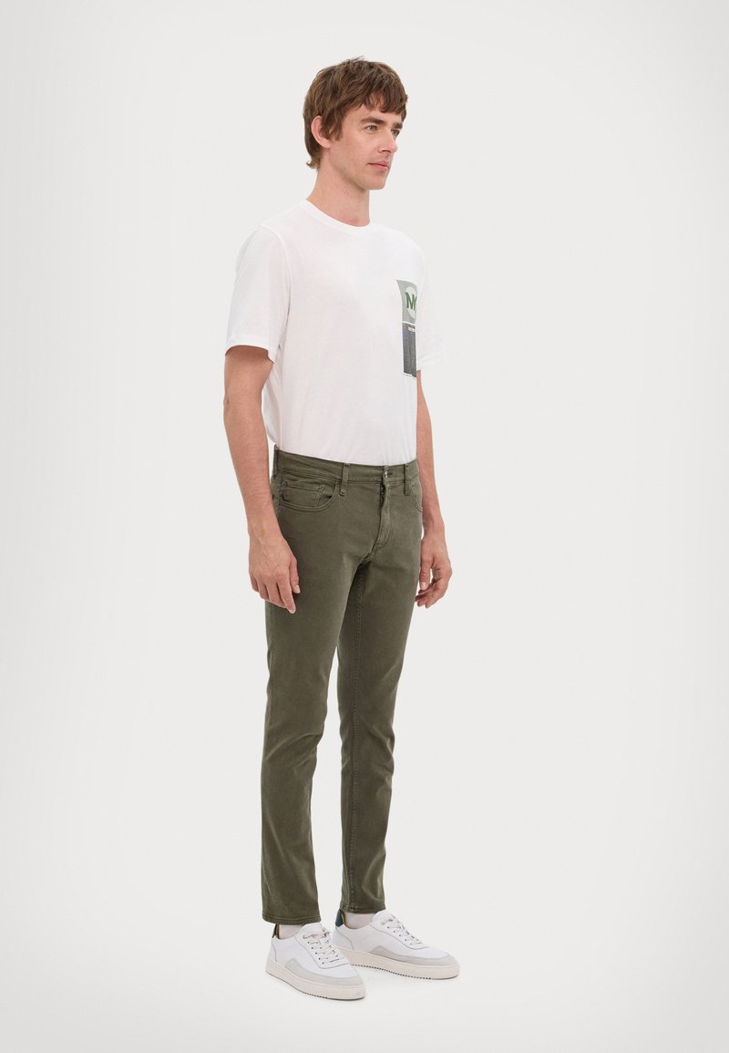 Fehér, rövid ujjú póló zöld folttal, olíva zöld, slim-fit nadrág és fehér sportcipő. Egyszerű dizájn, minta vagy kiegészítők nélkül.