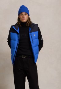 Zils pūkains bezmugurkaulnieks ar melnu apkakli un logo, uzvilkts pār melnā garajām piedurknēm. Modelis arī valkā zilu adītu cepuri un melnas bikses.