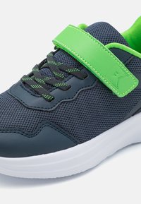 Sportcipő, amely navy színű hálós felsőrésszel, zöld bőr pánttal, fekete zsinórral zöld részletekkel és fehér gumitalppal rendelkezik.