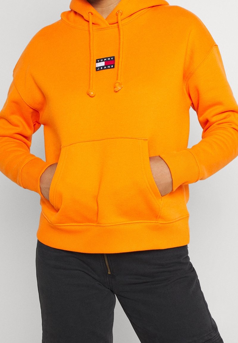 Sudadera con capucha naranja hecha de tela suave, con un bolsillo frontal, cordones ajustables y un pequeño parche con el logo "Tommy Jeans" en el pecho.
