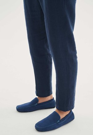 Jambes portant un pantalon slim bleu foncé associé à des mocassins en daim bleu assortis avec des coutures visibles sur fond blanc.