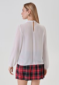 Weißes durchscheinendes Blusenshirt mit langen Ärmeln, dezenten Puff-Schultern und einem Schlüsselelement im Rücken, kombiniert mit einem schwarz-roten karierten Faltenrock.