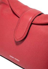 Rød læderhåndtaske med struktureret overflade, der har en klaplukning og en kontrasterende sort kant. Viser "GERRY WEBER" branding i hvid.