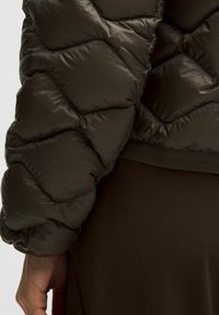 Zbliżenie na osobę ubraną w ciemnobrązową kurtkę puchową z pikowanym przeszyciem i pasującymi brązowymi spodniami, z widokiem na rękaw i dłoń.