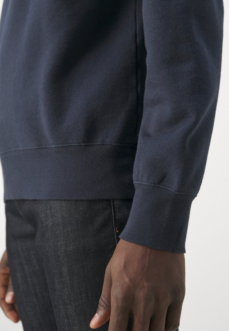 Dunkelhäutige Person, die einen navyblauen Langarmpullover und schwarze Denim-Jeans trägt, mit einer Nahaufnahme des Arms und der Hand.