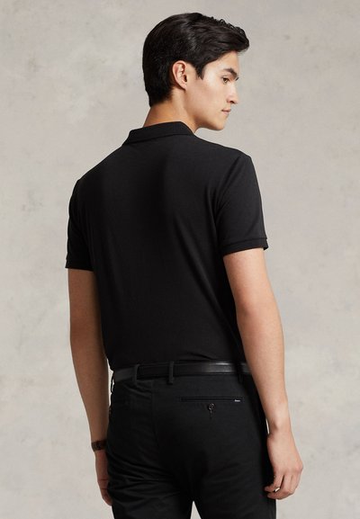 Junger Mann mit dunklen Haaren, der ein eng anliegendes schwarzes Poloshirt und schwarze Hose trägt, steht leicht seitlich gedreht vor einem einfarbigen Hintergrund.