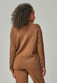 Brauner Pullover mit gerippter Textur und langen Ärmeln, ausgestattet mit einem abgerundeten Saum und seitlichen Schlitzen für mehr Komfort. Getragen mit passenden Hosen.