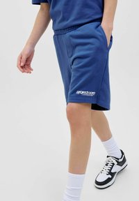 Blå sweatshirttygsbyxor med elastisk midja, sidofickor och vit logotyptryck vid fållen. I kombination med vita och svarta sneakers.
