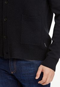 Marinblå cardigan i mjukt stickat tyg, med ribbad yta, knappfront och en enda ficka på vänster sida.