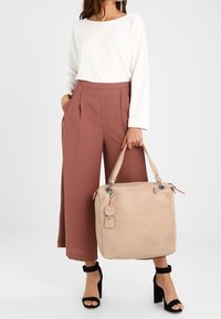 Beżowa torebka w kwadratowym kształcie, gładka skórzana tekstura, podwójne uchwyty i metalowe akcenty. W połączeniu z białą bluzką i różowymi spodniami typu wide-leg.