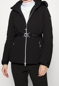 Black insulated jacket with a zippered front, white accents, textured sleeves, a belted waist, and a detachable hood featuring faux fur.