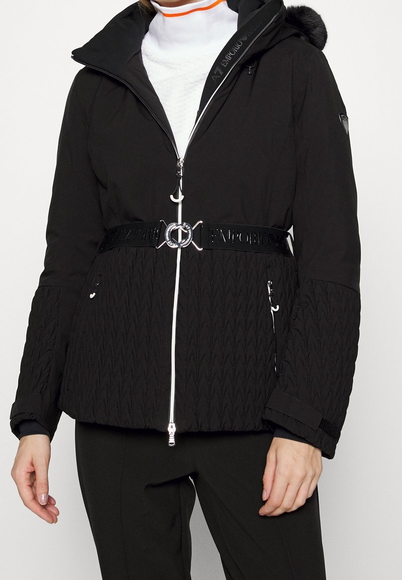 Black insulated jacket with a zippered front, white accents, textured sleeves, a belted waist, and a detachable hood featuring faux fur.