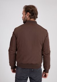 Man met oorbel die een bruine bomberjas en donkere spijkerbroek draagt, van achteren gezien tegen een effen achtergrond.