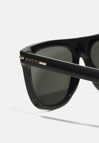 Melnas saulesbrilles ar tumšiem lēcu, ar biezu acetāta rāmi un zelta detaļām. "GUCCI" logo redzams sānā.