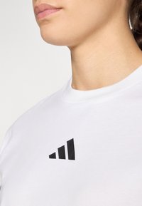 Bijela pamučna majica s crnim trokutastim grafičkim logotipom na lijevoj strani prsnog područja. Okrugli izrez sa glatkom teksturom.